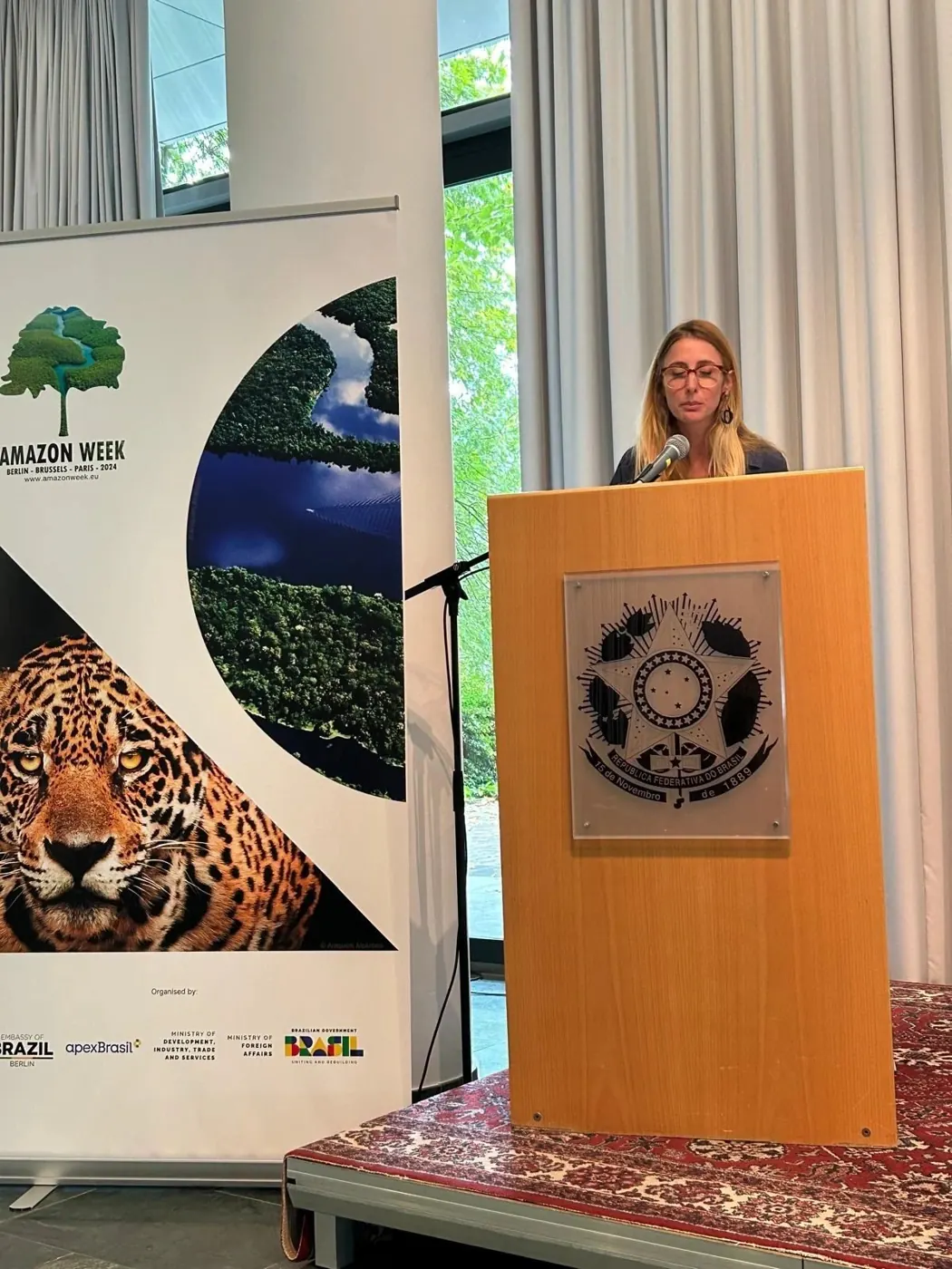 Priscila speaking at a lectern bearing the Brazilian coat of arms, next to an Amazon Week banner with a jaguar
